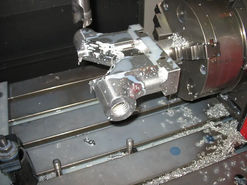 Complex milled metal component held in a CNC chuck with coolant on the bed